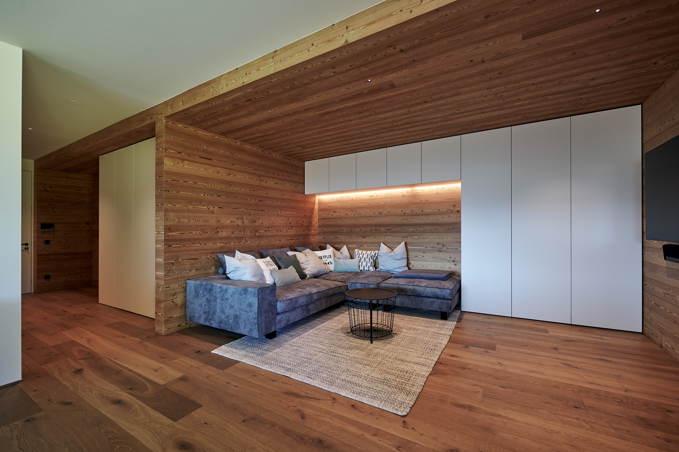 Gemütlicher Wohnbereich mit Wand- und Deckenverkleidung aus Mareiner Holz Piz Buin gebürstet. Die natürliche Holzoptik schafft ein warmes, einladendes Ambiente in Kombination mit modernen, weißen Einbauten und einer großen Couch.