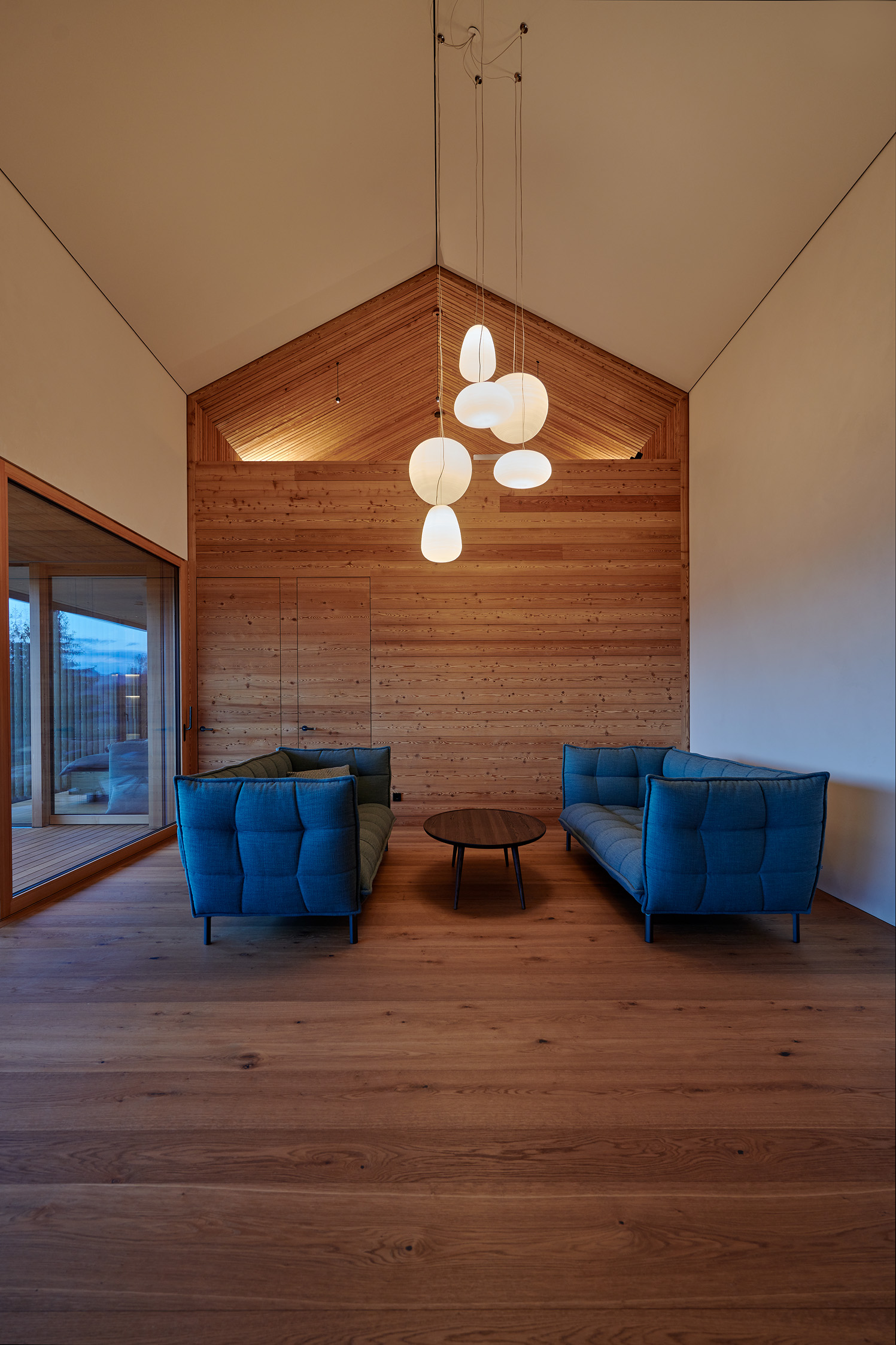 Wohnraum mit Holzverkleidung Alpine Wandpaneele Piz Buin gebürstet an Giebelseite, zwei blauen Sofas, runder Holztisch und modernen Hängeleuchten unter hoher Decke.
