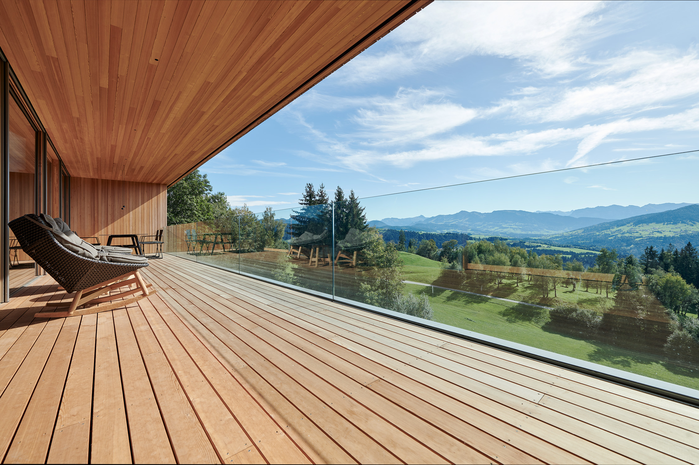 Große Holzterrasse mit Glasgeländer und atemberaubendem Ausblick ins Alpenvorland, Boden und Decke in Alpine Wandpaneele Piz Buin gebürstet, gemütlicher Liegestuhl im Vordergrund.