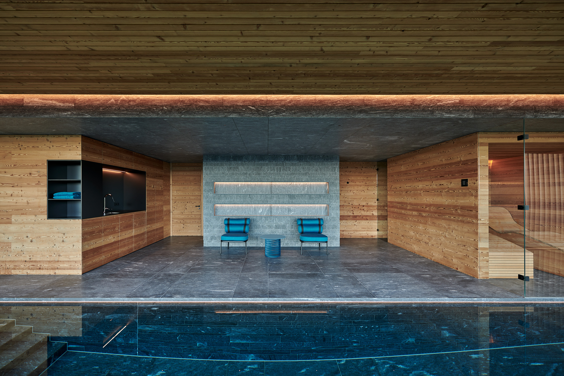 Moderner Wellnessbereich mit Pool, Holzverkleidung Alpine Wandpaneele Piz Buin gebürstet, grauen Steinfliesen, zwei blauen Sesseln vor Betonwand mit Lichtnischen.