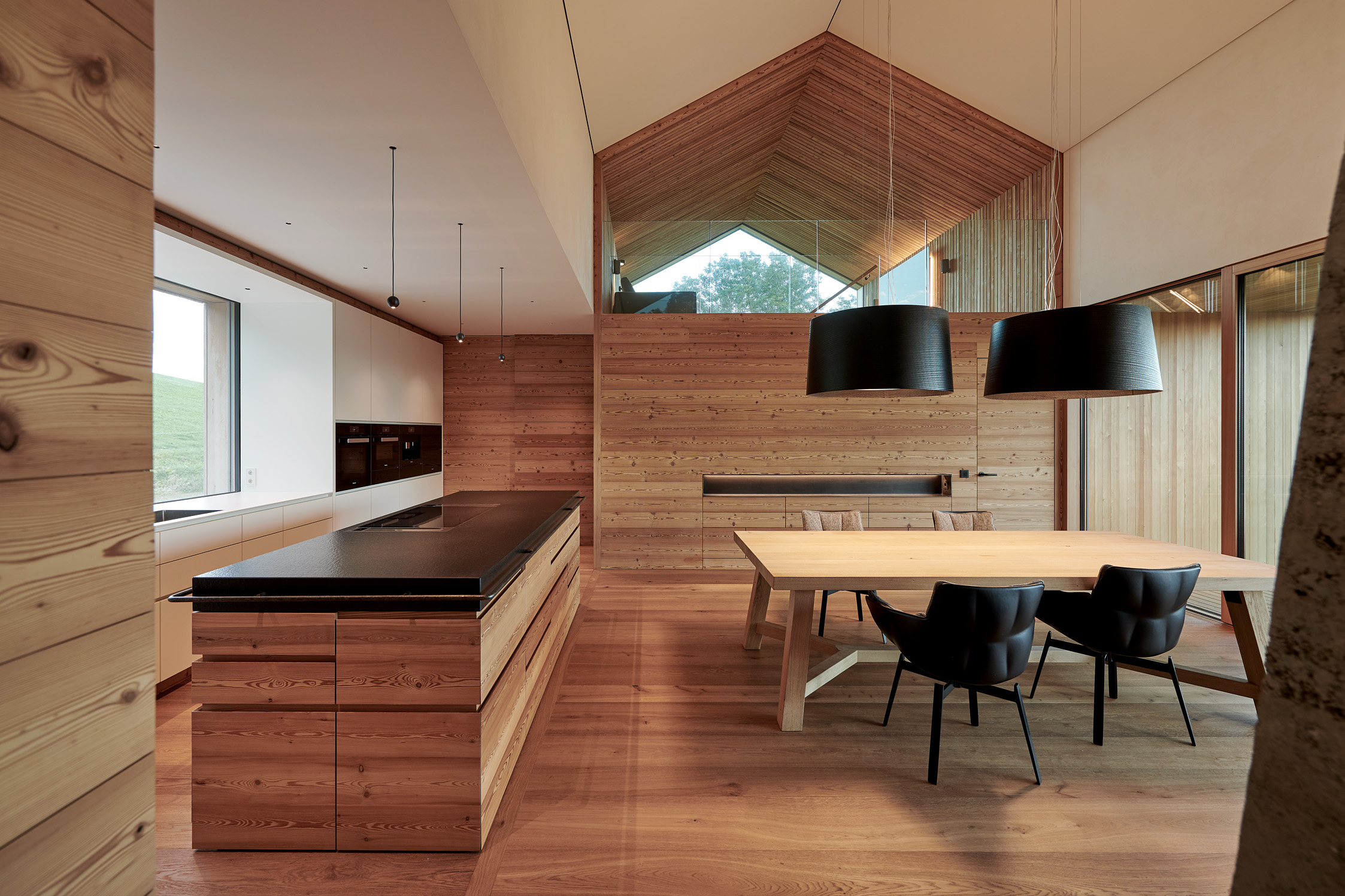 Moderne Wohnküche mit Holzverkleidung Alpine Wandpaneele Piz Buin gebürstet, Kochinsel mit schwarzer Arbeitsfläche, großem Holzesstisch und schwarzen Hängeleuchten unter hoher Decke.
