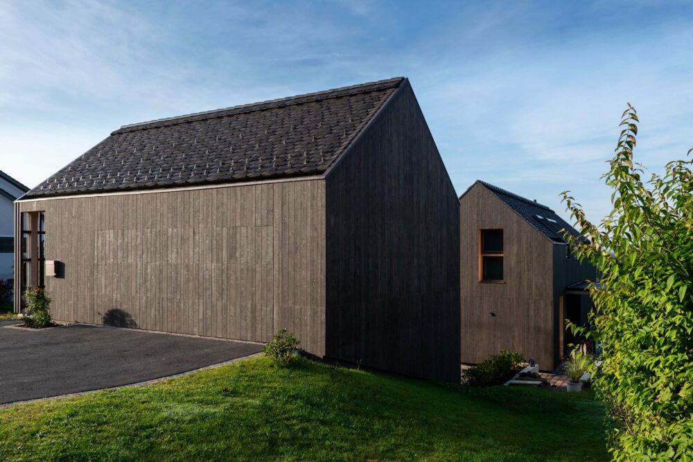 Zwei moderne Einfamilienhäuser mit dunkler, vertikaler Holzfassade stehen nebeneinander auf einem sanft abfallenden Grundstück. Die reduzierte Architektur mit klassischen Satteldächern wird durch die markante Struktur und natürliche Farbgebung der Holzfassade betont.