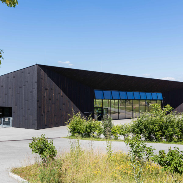 Modern, black building architecture with a striking, sloping roof shape and large glass front. The façade is made of dark charred wood (Shou Sugi Ban look) and forms a strong contrast to the surrounding green landscape. In the foreground is a path, wild flowers and a tree. The words "museumstrakt" can be seen on the building.