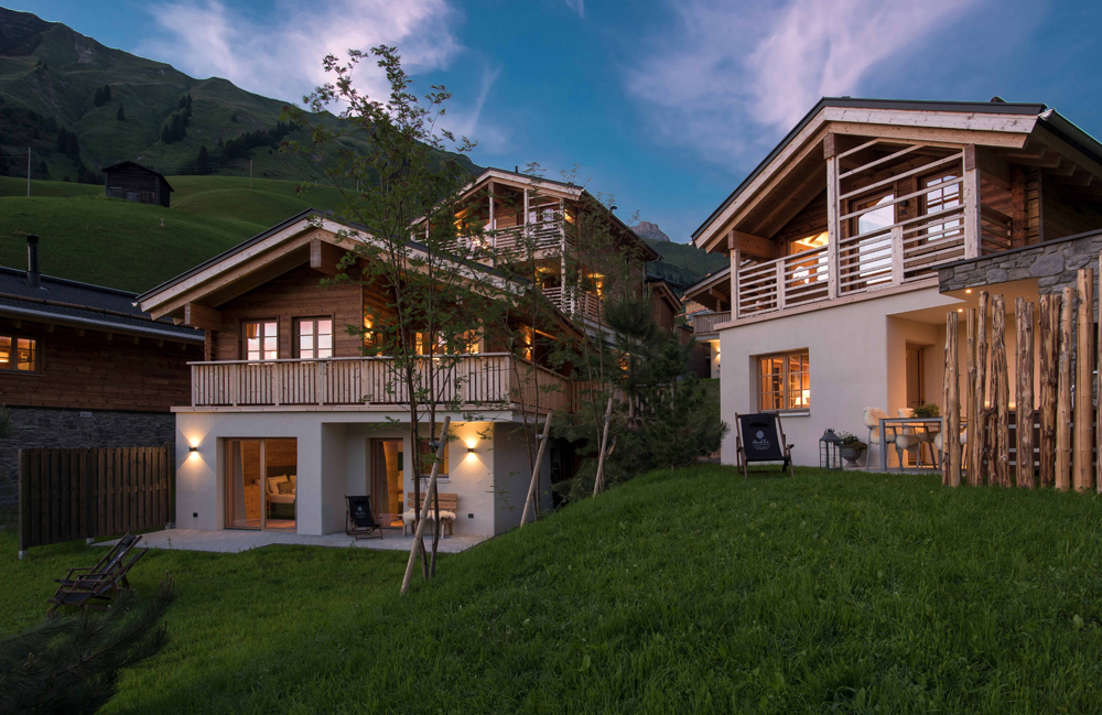 Atmospheric evening shot of the Aadla Walser chalets on an alpine slope. The facades of the chalets combine light-colored, plastered lower floors with rustic wood on the upper floors, made from the products Piz Bernina Lava and Marmolada. Spacious balconies, wooden elements and warm lighting create a cozy atmosphere.