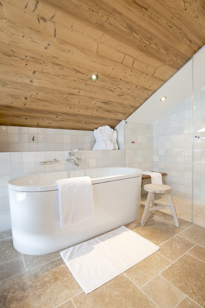 Modernes Badezimmer in der Lech Lodge mit freistehender Badewanne, hellen Fliesen und Dachschräge aus alpiner Wandpaneele „Gran Paradiso“. Die natürliche Holzdecke sorgt für Wärme und Gemütlichkeit im eleganten Wellnessambiente.