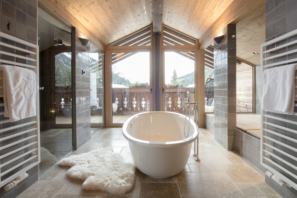 Luxuriöses Badezimmer in der Lech Lodge mit freistehender Badewanne, bodentiefen Fenstern und alpiner Wandpaneele „Gran Paradiso“. Edle Materialien, Bergblick und warme Holzdetails schaffen ein exklusives Spa-Erlebnis im Chalet-Stil.