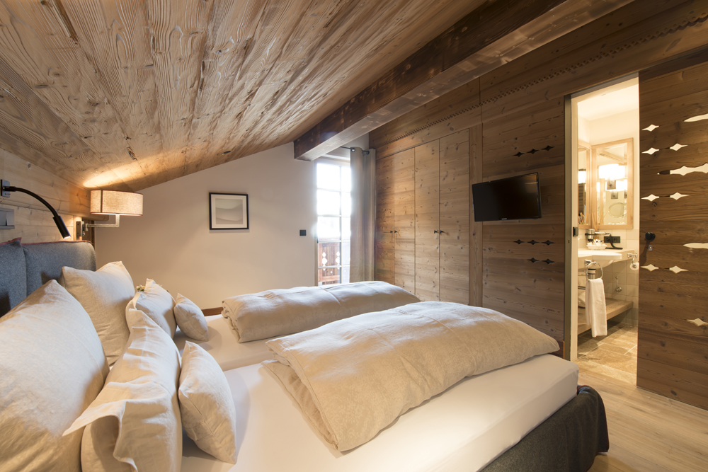 Stilvolles Chalet-Schlafzimmer in der Lech Lodge, gestaltet mit der alpinen Wandpaneele „Mont Blanc“. Warmes, rustikales Holz mit traditionellen Ausstanzungen verleiht dem Raum eine gemütliche Atmosphäre im gehobenen Alpenstil.