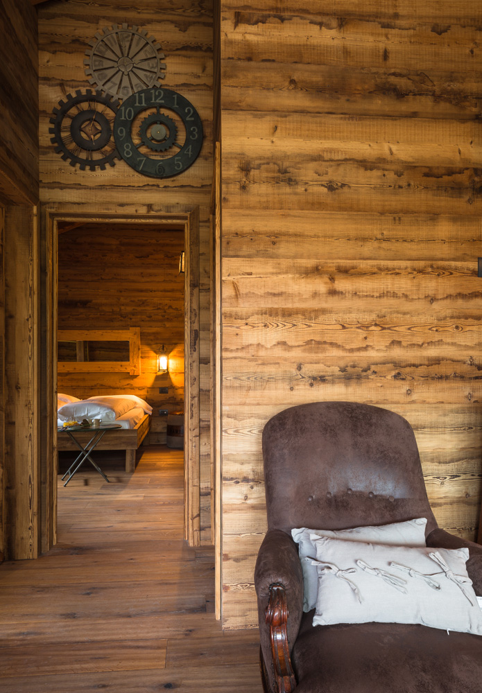 Gemütliches Interieur mit Wand- und Bodenverkleidung aus AWP Piz Badile Holz, Blick in ein Schlafzimmer mit Holzmöbeln und warmem Licht, im Vordergrund ein brauner Sessel mit Kissen.