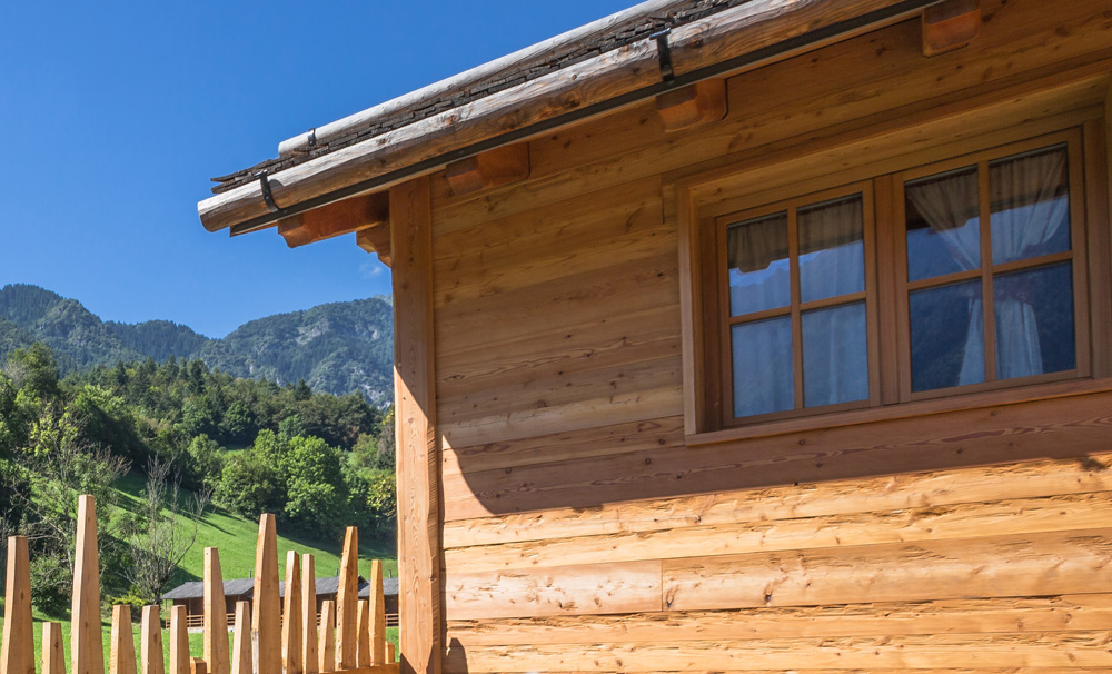 Detailaufnahme einer sonnenbeschienenen Holzfassade aus Alpine Wandpaneele Piz Bernina mit Fenster und traditioneller Dachkonstruktion vor alpiner Landschaft.