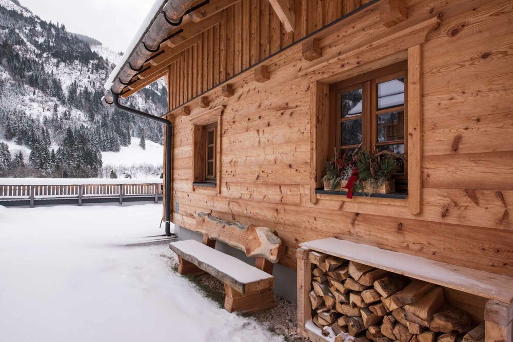 Fassade einer verschneiten Berghütte mit Alpine Wandpaneelen Piz Buin in natürlichem Holzton, rustikaler Oberfläche und Fensterdekoration im Bergdorf Donnersbachwald.
