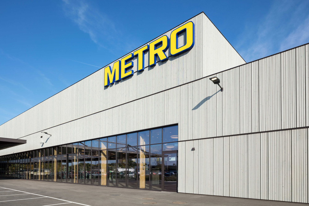 METRO-Gebäude in St. Pölten mit moderner Holzfassade aus vertikalen Elementen in Thermofichte vorvergraut, darüber gelb-blaues METRO-Logo, klarer blauer Himmel im Hintergrund.