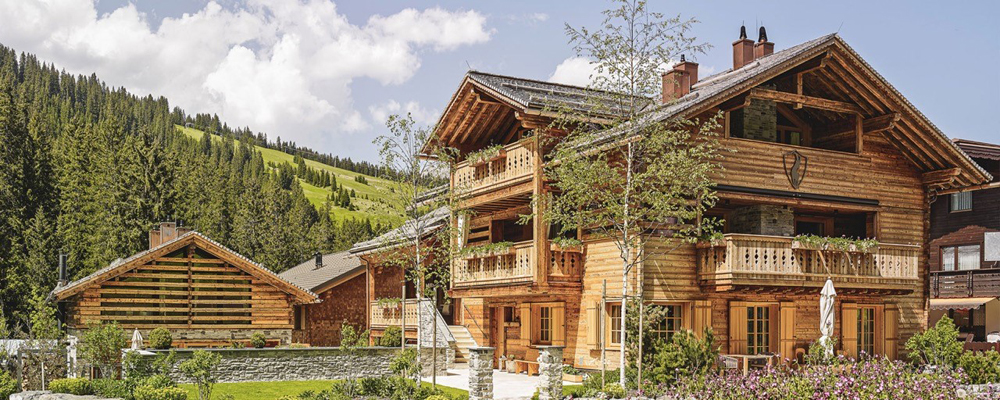 Außenansicht der Lech Lodge im Sommer mit Fassade aus alpiner Wandpaneele „Piz Bernina Lava“. Traditionelles Chaletdesign trifft auf moderne Holzarchitektur inmitten blühender Landschaft und grüner Berghänge.