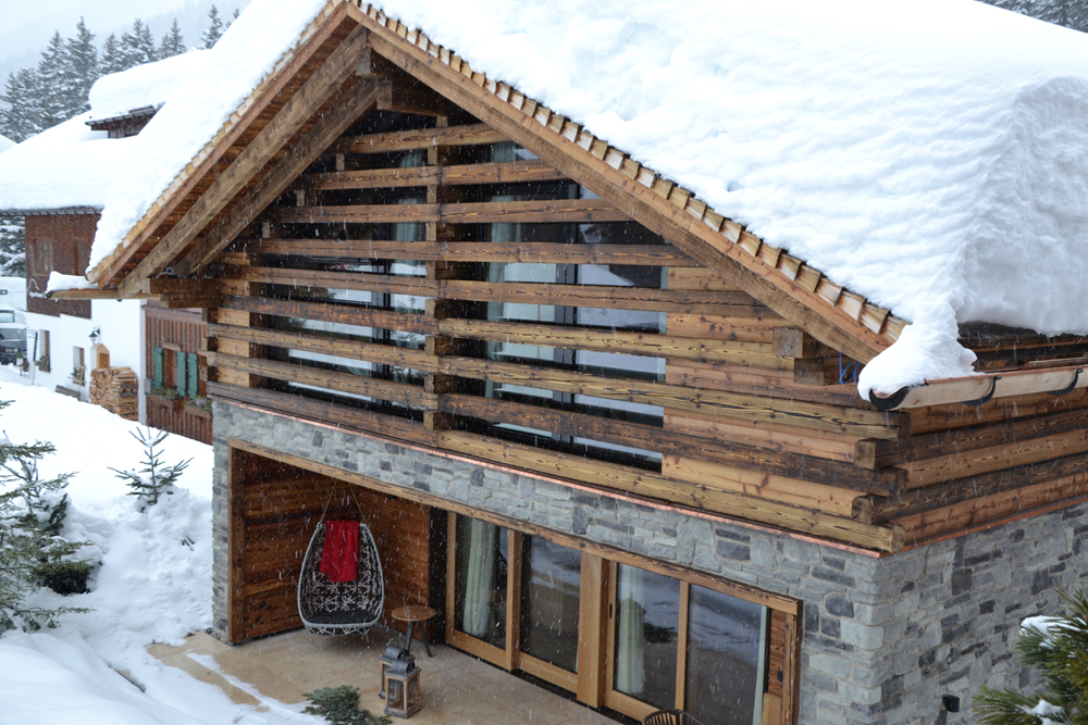 Außenansicht der Lech Lodge mit charaktervoller Fassade aus alpiner Wandpaneele „Piz Bernina Lava“. Die rustikale Holzverkleidung im Wechselspiel mit Naturstein und großen Glasflächen vermittelt modernes Alpenflair im tief verschneiten Bergdorf.