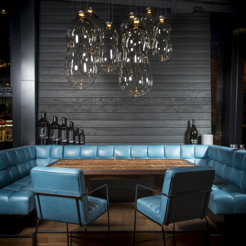 Innenansicht der Spelunke Bar in Wien mit alpinen Wandpaneelen Vulcano in dunkler, verkohlter Optik. Die Wandverkleidung aus horizontalen Holzelementen bildet den Hintergrund für einen massiven Holztisch, umgeben von türkisfarben gepolsterten Sitzbänken und Sesseln. Auffällige Glaspendel-Leuchten hängen über dem Tisch und sorgen für ein elegantes, modernes Ambiente.