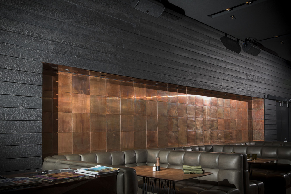 Dunkle Wand mit alpinen Wandpaneelen Vulcano im Restaurant Spelunke in Wien. Die schwarze, strukturierte Holzverkleidung umrahmt eine kupferverkleidete Nische mit integrierter Beleuchtung. Davor befinden sich braune Lederbänke und Holztische in stilvoller Lounge-Atmosphäre.