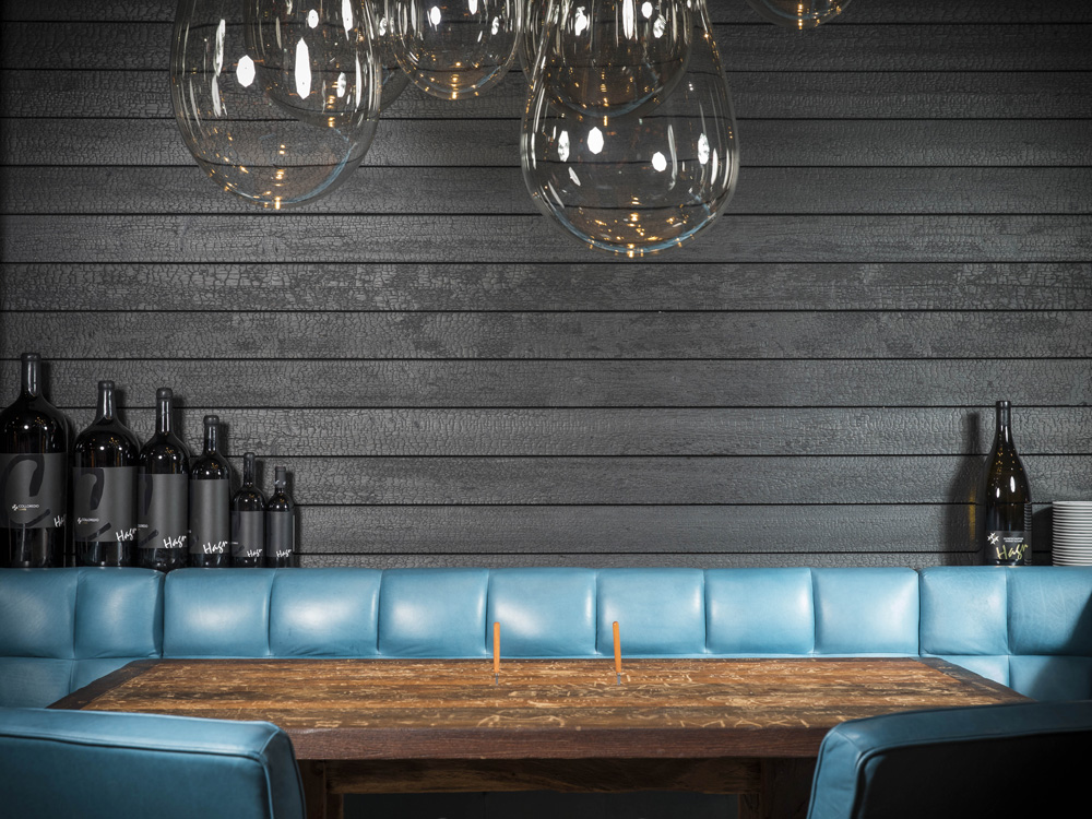 Interior shot from the Spelunke restaurant in Vienna with elegant wall design made of Alpine wall panels Vulcano. The black charred wood surface in a shou-sugi-ban look creates a stylish and contrasting atmosphere in combination with the turquoise-coloured bench, the rustic wooden table and modern glass body lamps.
