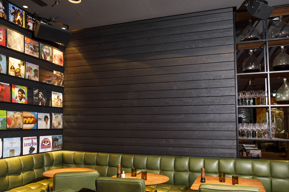 Innenaufnahme des Restaurants Spelunke in Wien mit Wandverkleidung aus Alpine Wandpaneele Vulcano. Die verkohlte Holzstruktur im Shou-Sugi-Ban-Stil verleiht dem Raum eine warme, dunkle Eleganz, ergänzt durch grüne Polsterbänke, runde Holztische, ein Weinregal und eine kreative Wandgestaltung mit Plattencovern.