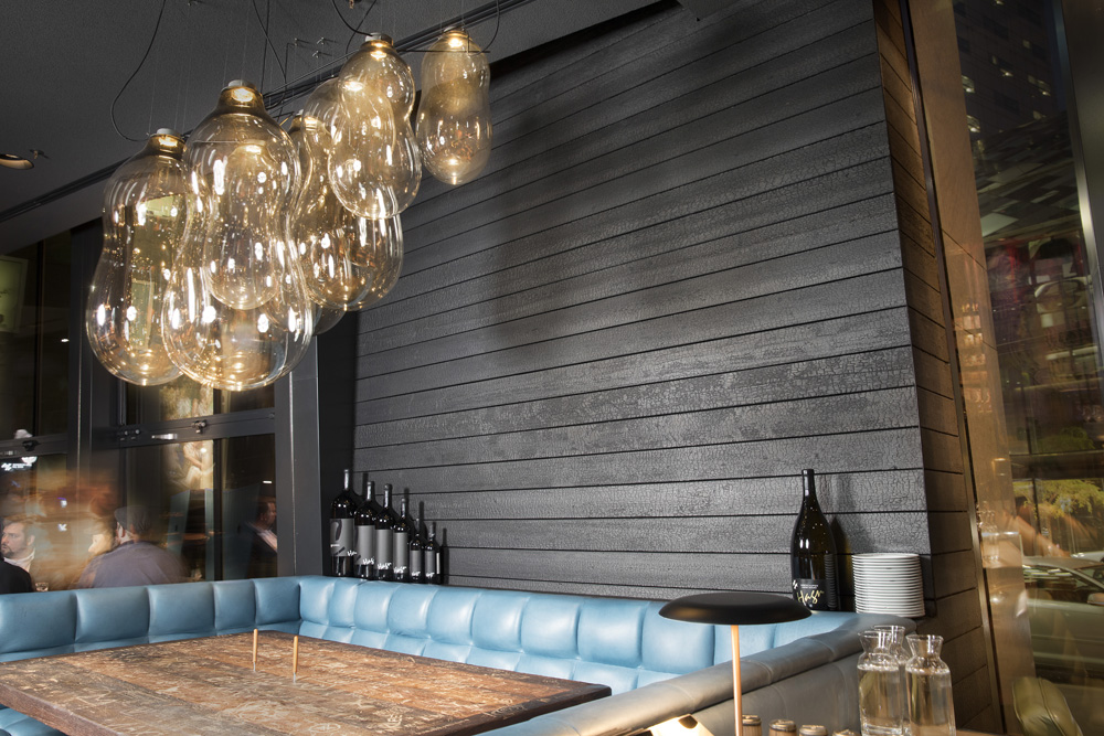 Restaurantbereich in der Spelunke Wien mit einer Wand aus Alpine Wandpaneele Vulcano in verkohlter Holzoptik. Im Vordergrund befindet sich eine elegante Sitzecke mit türkisfarbenen Polstern, ein massiver Holztisch und eine markante Leuchte aus mundgeblasenen Glaslampen. Das moderne Ambiente wird durch die dunkle Holzstruktur stilvoll unterstrichen.