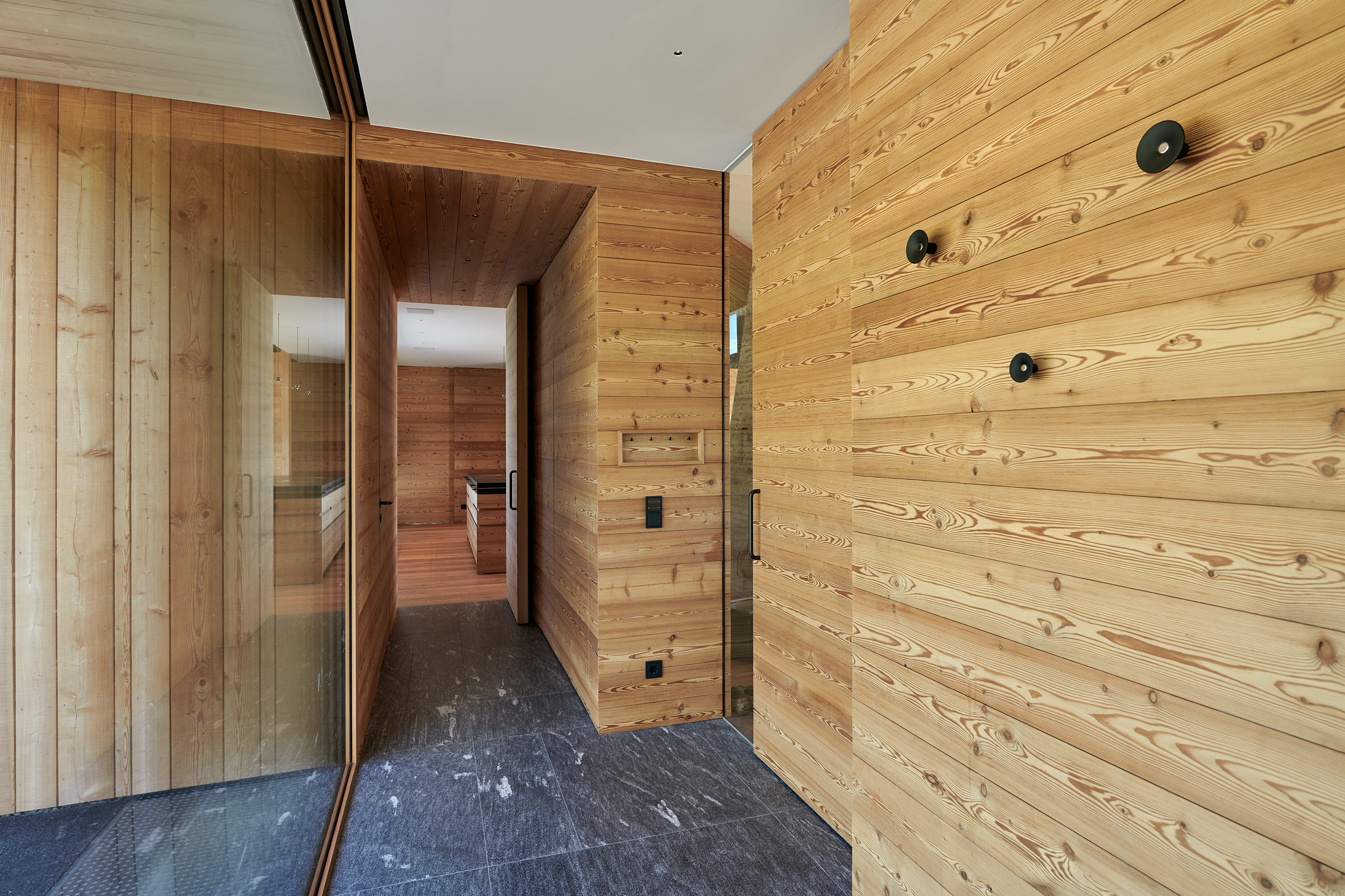Eingangsbereich mit gebürsteter Alpine Wandpaneele Piz Buin Wandverkleidung aus naturbelassenem Holz. Die helle Holzmaserung setzt einen warmen Kontrast zum dunklen Natursteinboden. Glaselemente sorgen für transparente Übergänge zwischen den Räumen und unterstreichen das moderne, reduzierte Designkonzept.