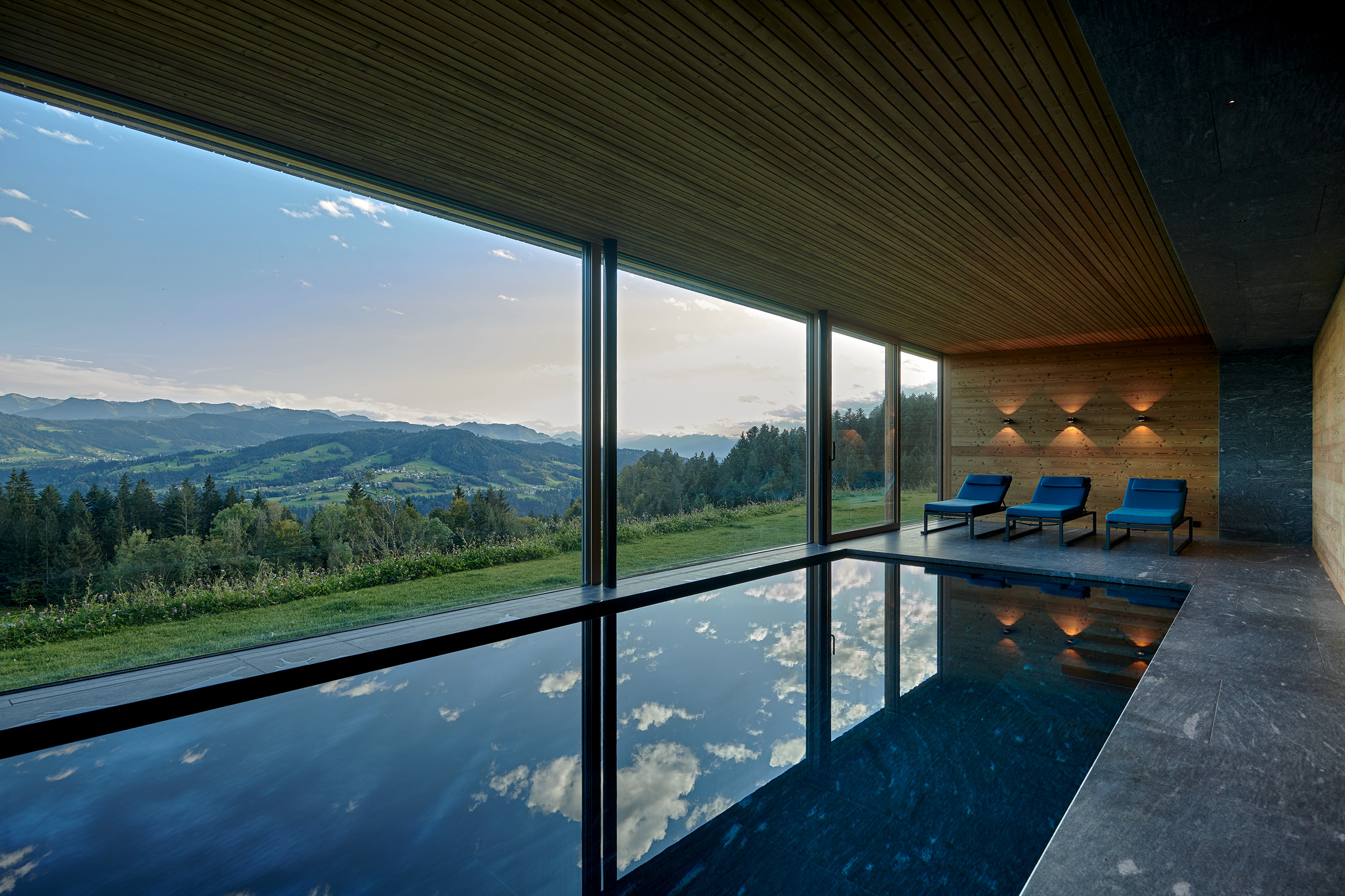 Hochwertig gestalteter Indoor-Poolbereich mit Panoramaverglasung und Blick auf die hügelige Landschaft. Die Decke und Wandverkleidung aus gebürsteten Alpine Wandpaneele Piz Buin Paneelen sorgt für eine warme, natürliche Atmosphäre. Blaue Liegen und gezielte Beleuchtung runden das stilvolle Design ab.