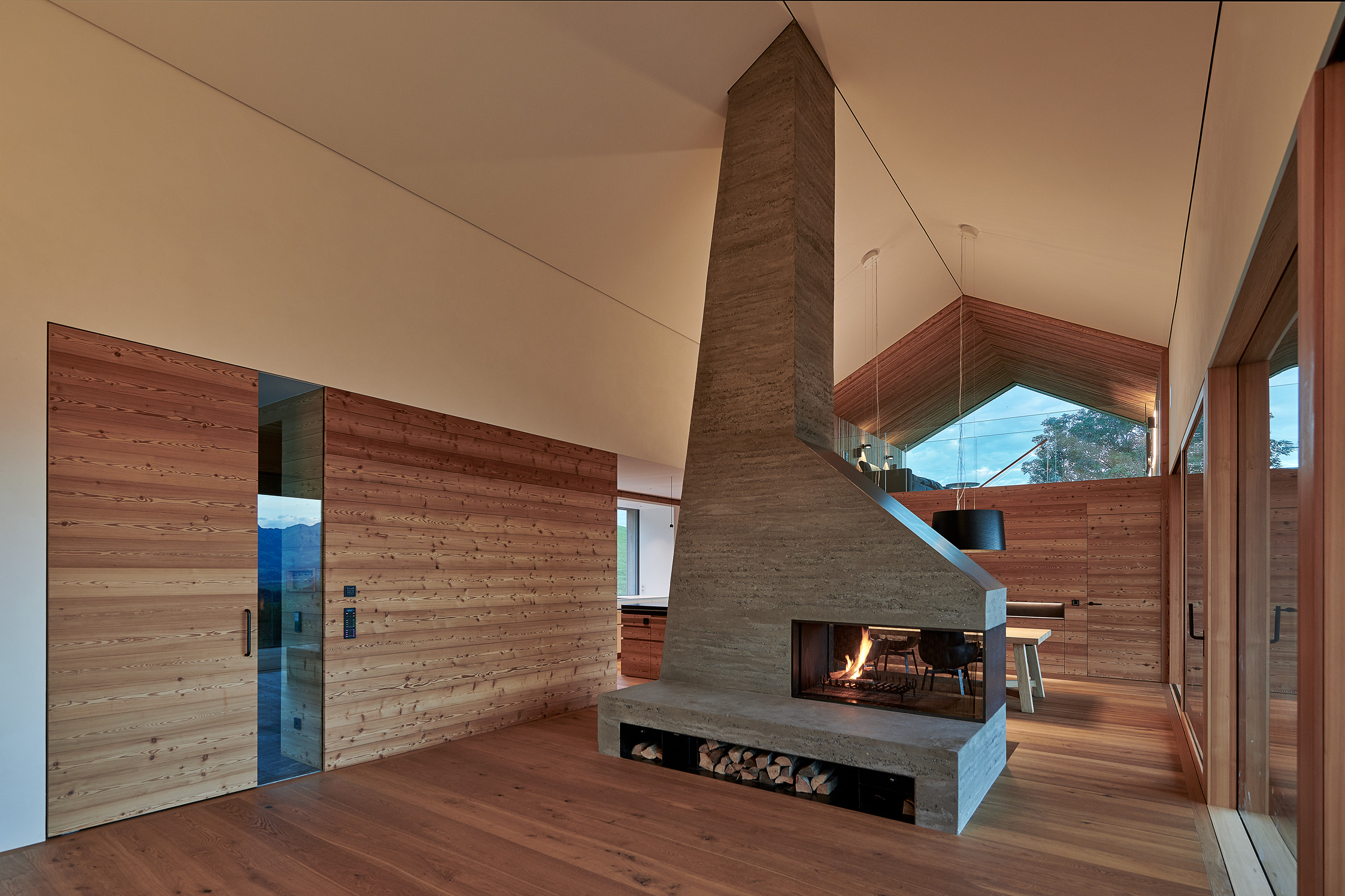 Wohnbereich mit gebürsteter Alpine Wandpaneele Piz Buin Wand- und Deckenverkleidung in naturbelassenem Holzton. Ein markanter Kamin aus Sichtbeton steht im Zentrum des Raumes, ergänzt durch großflächige Fensterfronten, die den Blick auf die Berglandschaft freigeben.