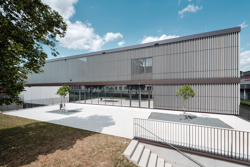 Moderne Schulgebäude-Fassade der HTL Zistersdorf mit vertikalen Lamellen aus vorvergrauter Thermofichte, symmetrisch gestaltet mit großen Glasflächen und zwei kleinen Bäumen im Vorplatz.