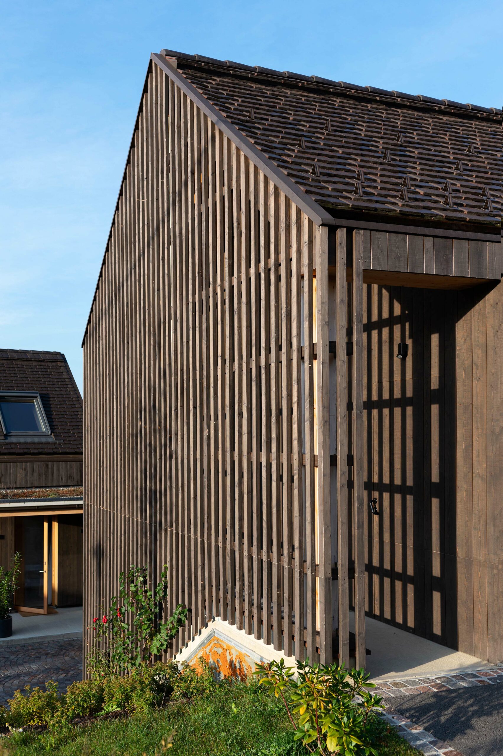 Detailansicht eines modernen Wohnhauses mit vertikaler Holzlattung aus Mareiner Holz. Die vertikale Struktur der Fassade erzeugt ein markantes Schattenspiel auf der darunterliegenden dunklen Holzverkleidung. Der Giebelbau mit Ziegeldach steht vor blauem Himmel, flankiert von einem kleinen Gartenbereich mit Bepflanzung im Vordergrund.