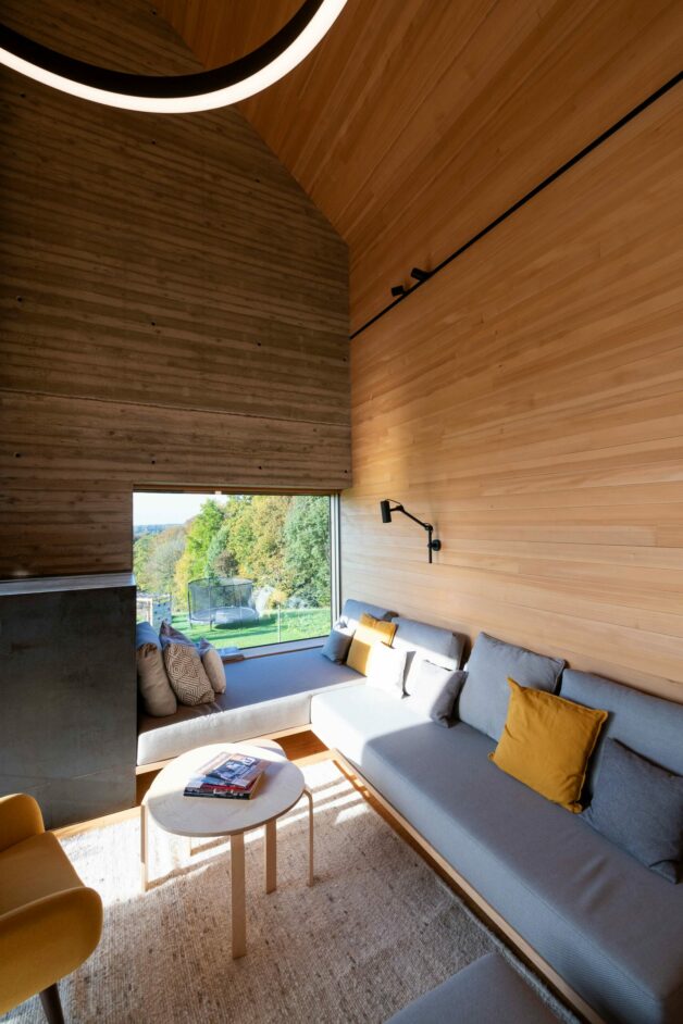 Wohnraum im Privathaus Hengsberg mit hoher Decke und großem Panoramafenster. Die Innenwände sind mit hellem Holz verkleidet und verleihen dem Raum eine warme, natürliche Atmosphäre. Ein graues Sofa mit gelben Kissen und moderne Leuchten ergänzen das reduzierte, stilvolle Design.