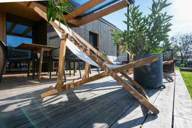 Sonnige Holzterrasse des Privathauses Hengsberg mit Liegestuhl und Sitzbereich. Die Terrasse ist ergänzt durch Pflanzen in modernen Töpfen und eine Überdachung aus Glas und Holz, die für ein angenehmes Outdoor-Ambiente sorgt.