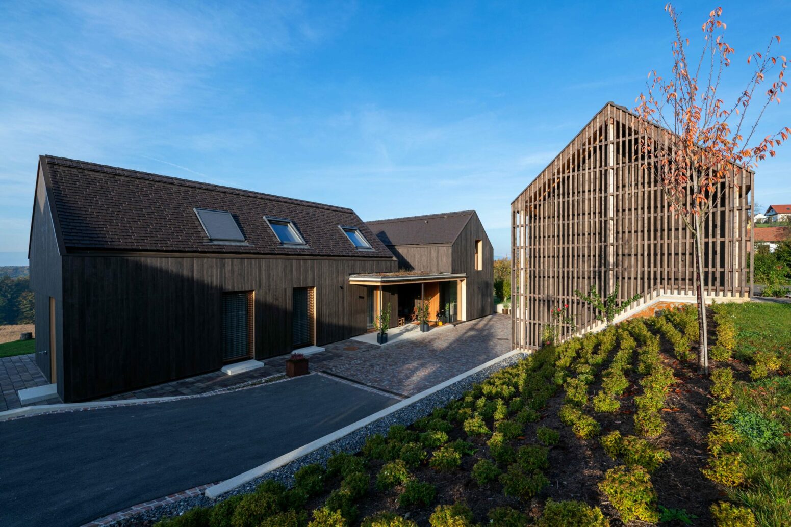 Modernes Wohnhaus-Ensemble in Hengsberg mit vertikaler und horizontaler Holzfassade von Mareiner Holz. Die dunkle, strukturierte Holzverkleidung betont die klare Formensprache der Architektur und harmoniert mit den begrünten Außenbereichen und Natursteinwegen.