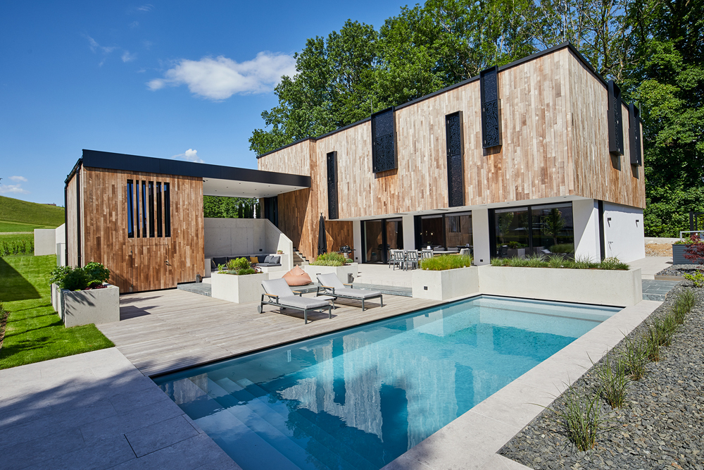 Modernes Einfamilienhaus mit vertikal strukturierter Holzfassade von Mareiner Holz, gestaltet von raumbau architekten Salzburg. Große Fensterflächen, Poolbereich und stilvolle Außenanlagen bei strahlendem Sonnenschein.