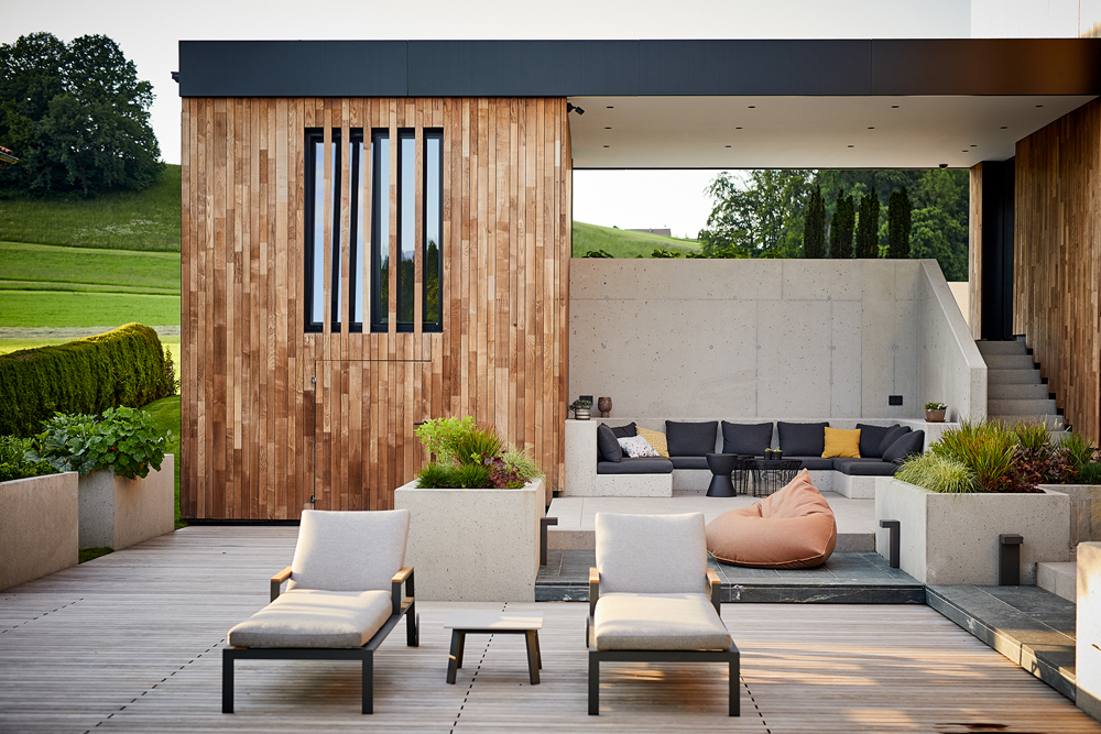Moderner Terrassenbereich eines Privathauses mit vertikaler Holzfassade aus Mareiner Holz. Im Vordergrund stehen zwei Liegestühle und ein kleiner Tisch auf einem Holzdeck. Dahinter gemütlicher Lounge-Bereich mit Sitzpolstern und Betonwänden, seitlich eine Außentreppe.