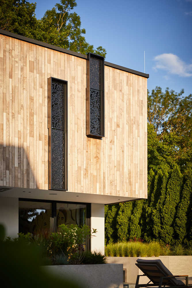 Nahaufnahme der modernen Holzfassade aus vertikalen Gardasee-Paneelen am Privathaus Blaschke, gestaltet von raumbau architekten. Schwarze vertikale Fensterrahmenelemente setzen stilvolle Akzente in der hellen Holzoberfläche, eingerahmt von dichter Vegetation.