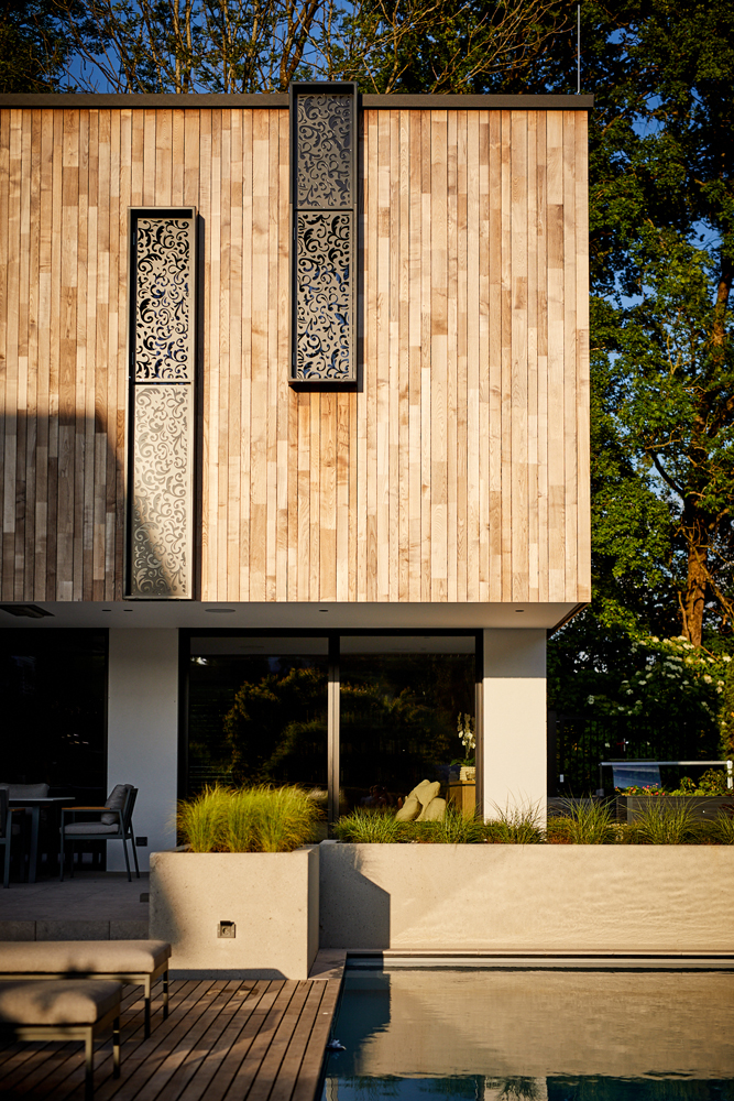 Nahaufnahme der oberen Etage eines modernen Wohnhauses mit vertikaler Holzfassade aus Mareiner Holz. Zwei schmale, dekorative Fenster mit schwarzem Ornamentrahmen setzen Akzente. Darunter große Glasfront mit Blick auf Terrasse und Pool.