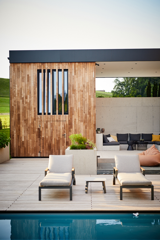 Detailansicht des Poolbereichs eines modernen Hauses mit vertikal verbauter Holzfassade aus Mareiner Holz. Zwei Sonnenliegen stehen auf der Holzterrasse vor einem Lounge-Bereich mit Sichtbetonwand und stilvoller Überdachung.