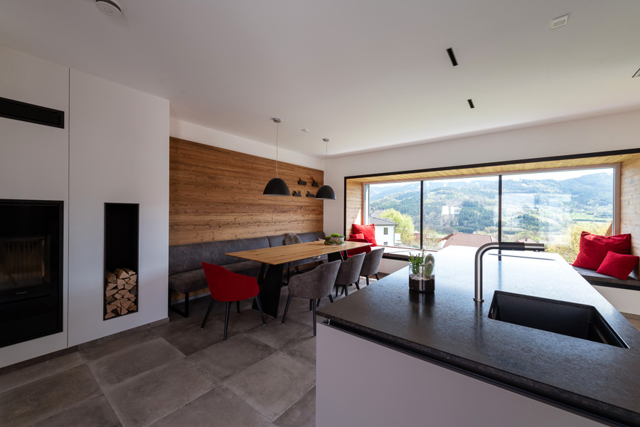 Modern gestalteter Wohn-Essbereich in einem Privathaus mit großer Fensterfront und Panoramablick ins Grüne. Die Rückwand des Essbereichs ist mit warmem Holz – Gran Paradiso – gestaltet, ergänzt durch eine gemütliche Eckbank und moderne Hängelampen. In der Fensterlaibung ist eine gemütliche Sitznische mit roten Kissen integriert.