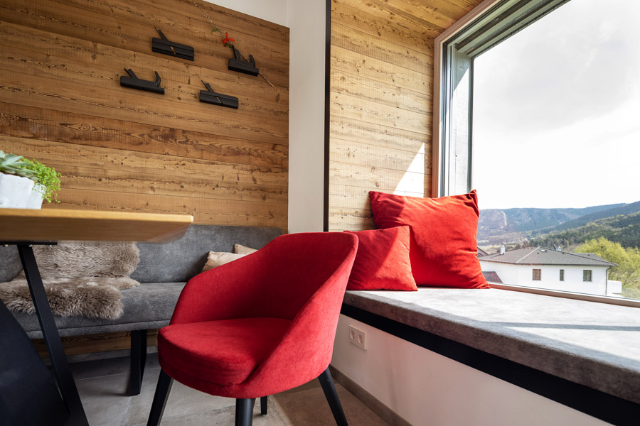 Gemütliche Sitzecke in einem modern eingerichteten Wohnraum mit rustikaler Holzverkleidung – Gran Paradiso – an der Wand. Die große Fensterlaibung ist mit grauem Polster und leuchtend roten Kissen ausgestattet und bietet einen Ausblick auf eine hügelige Landschaft. Eine rote Polsterstuhl setzt einen farblichen Akzent.