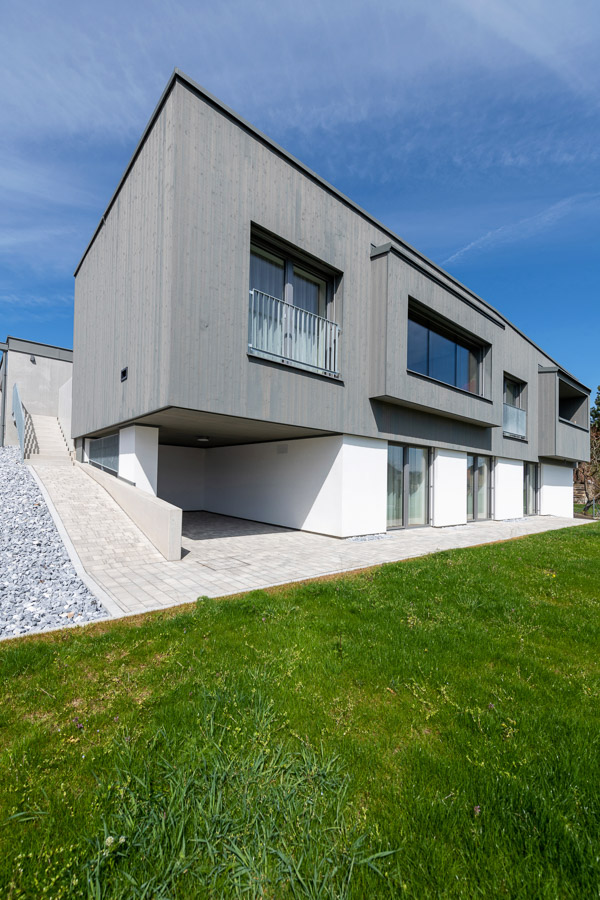 Modernes Einfamilienhaus mit klarer Linienführung und einer Fassade aus Gran Paradiso 212° Holz mit Bandsägeschnitt, vorvergraut. Die obere Etage ist mit grauer Holzverschalung verkleidet, während das Erdgeschoss in weißem Putz gehalten ist. Eine Rampe führt zur oberen Etage, das Haus steht auf einem gepflegten Grundstück mit grünem Rasen.