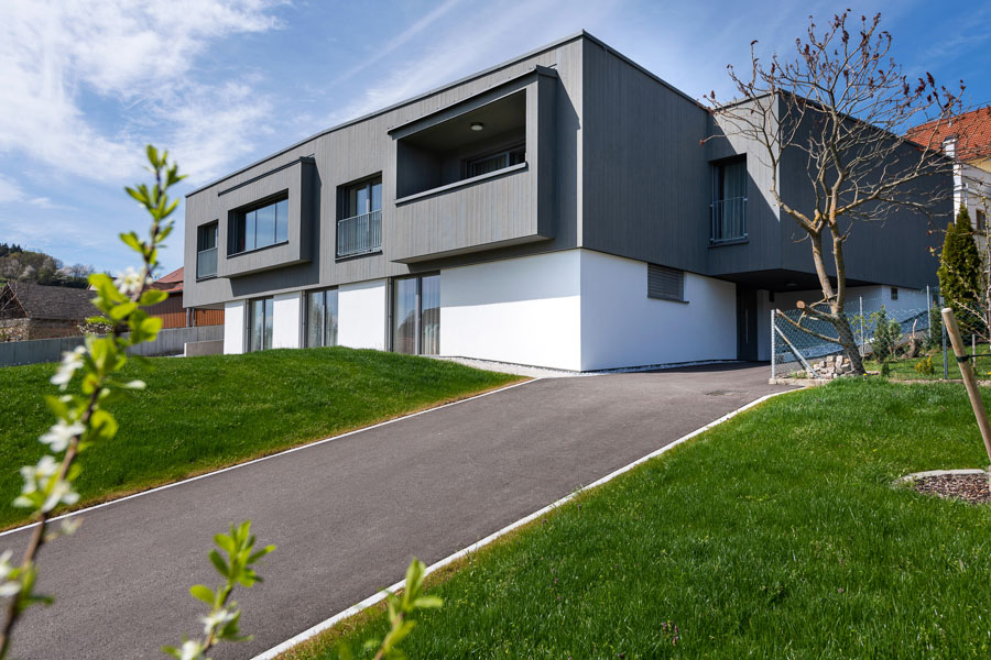 Modernes Einfamilienhaus mit klarer, kubischer Architektur. Die Fassade im Obergeschoss ist mit vertikalen Holzpaneelen des Produkts „Gran Paradiso 212°“ mit Bandsägeschnitt und Vorvergrauung gestaltet, das Erdgeschoss ist weiß verputzt. Eine asphaltierte Auffahrt führt zur überdachten Hauseingangszone.
