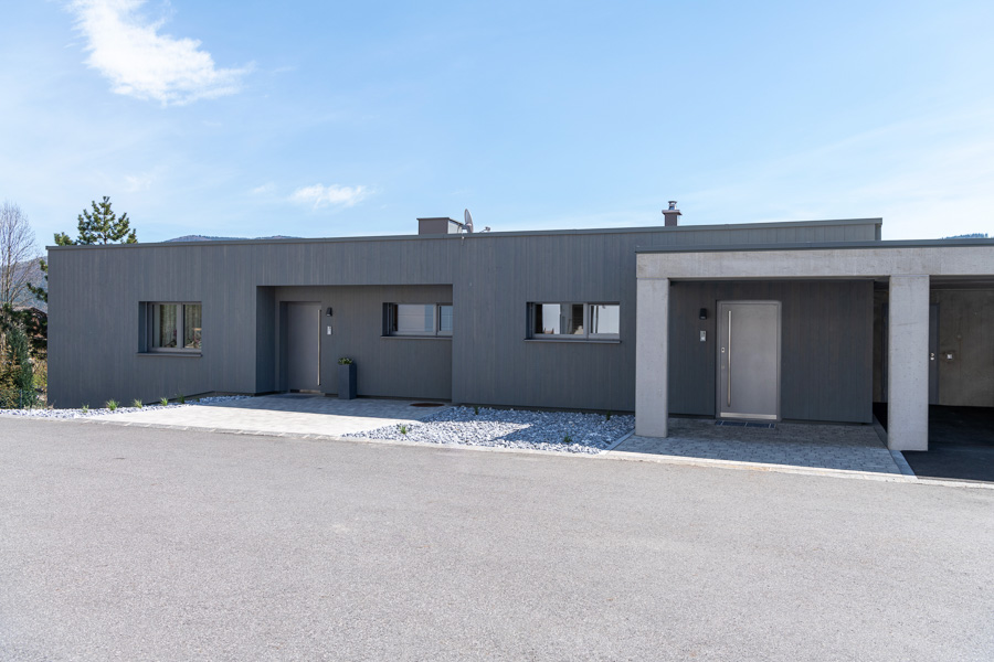 Straßenseitige Ansicht eines modernen, eingeschossigen Wohnhauses mit flacher Dachform. Die Fassade ist mit vertikalen Holzpaneelen in Grau gestaltet, ergänzt durch Sichtbetonelemente im Eingangs- und Carportbereich. Das Produkt an der Fassade ist „Gran Paradiso 212° mit Bandsägeschnitt & vorvergraut“.