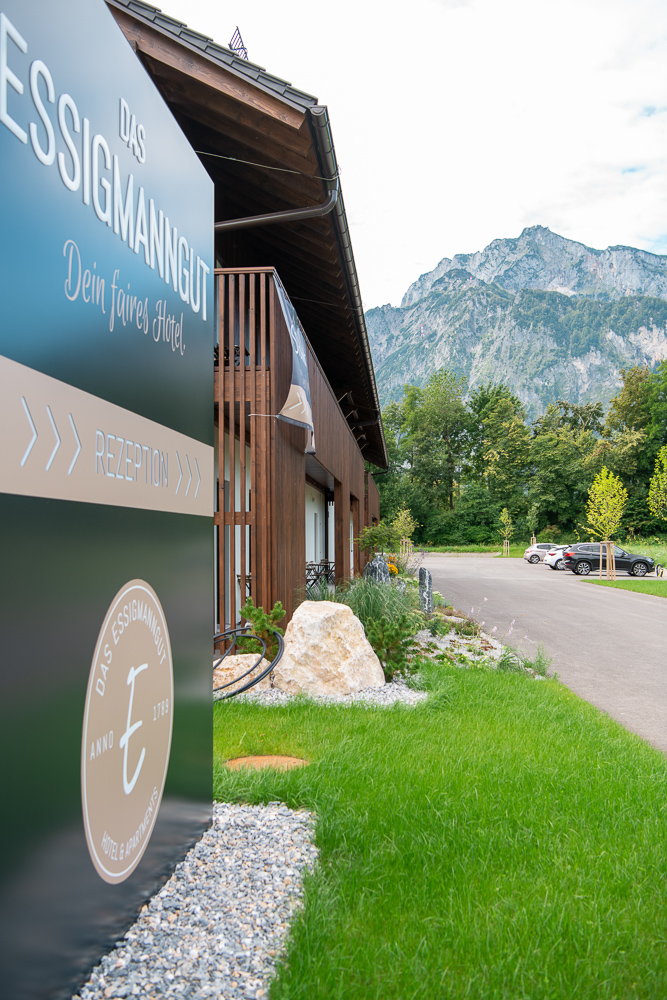 Eingangsbereich des Hotels „Das Essigmanngut“ in Salzburg mit modernem Empfangsschild im Vordergrund. Dahinter das Hotelgebäude mit vertikaler Holzfassade aus Mareiner Holz und beeindruckendem Bergpanorama im Hintergrund.