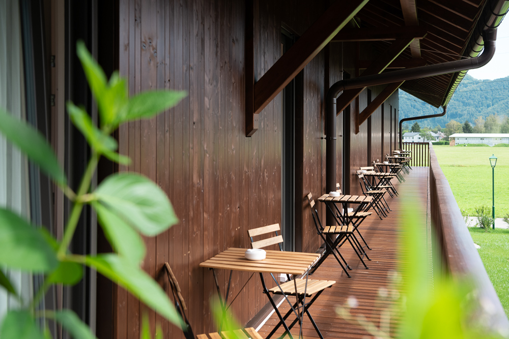 Balkonansicht des Hotels Essigmanngut in Salzburg mit blick auf eine Holzfassade. Entlang des Balkons stehen kleine Holzsitzgruppen vor den Gästezimmern. Im Hintergrund weite grüne Wiesen und bewaldete Hügel.