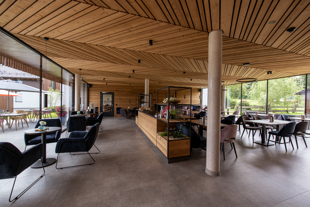 Moderner Hotel-Restaurantbereich mit decken- und wandverkleidetem Gran Paradiso 212° Holz von Mareiner. Große Glasfronten bieten Blick ins Grüne. Elegante Möblierung mit schwarzen Sesseln, rosa Stühlen und zentralem Regalmodul.