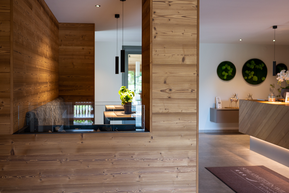 Helle Hotel-Lounge mit Wänden aus Mareiner Gran Paradiso 212° Holz. Im Vordergrund ein Glas-Kamin, dahinter eine Sitznische mit Tisch und Pflanze. Rechts moderne Rezeption mit runder Wanddeko und Hängeleuchten.
