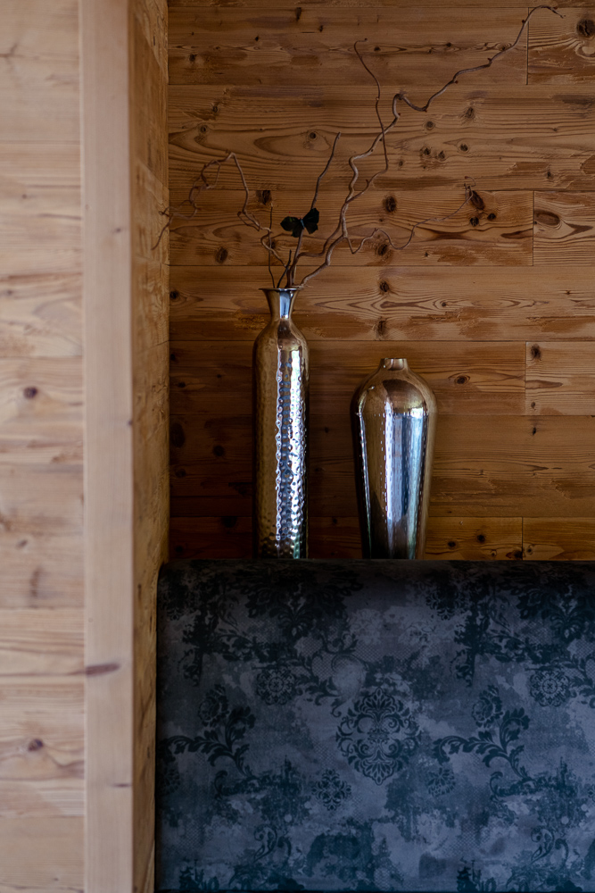 Detailansicht einer Sitznische im Hotel Essigmanngut mit Wandverkleidung aus Mareiner Mont Blanc Alpine Wandpaneelen. Zwei metallische Vasen mit Zweigen stehen dekorativ auf der Rückenlehne einer gepolsterten Bank mit floralen Mustern.
