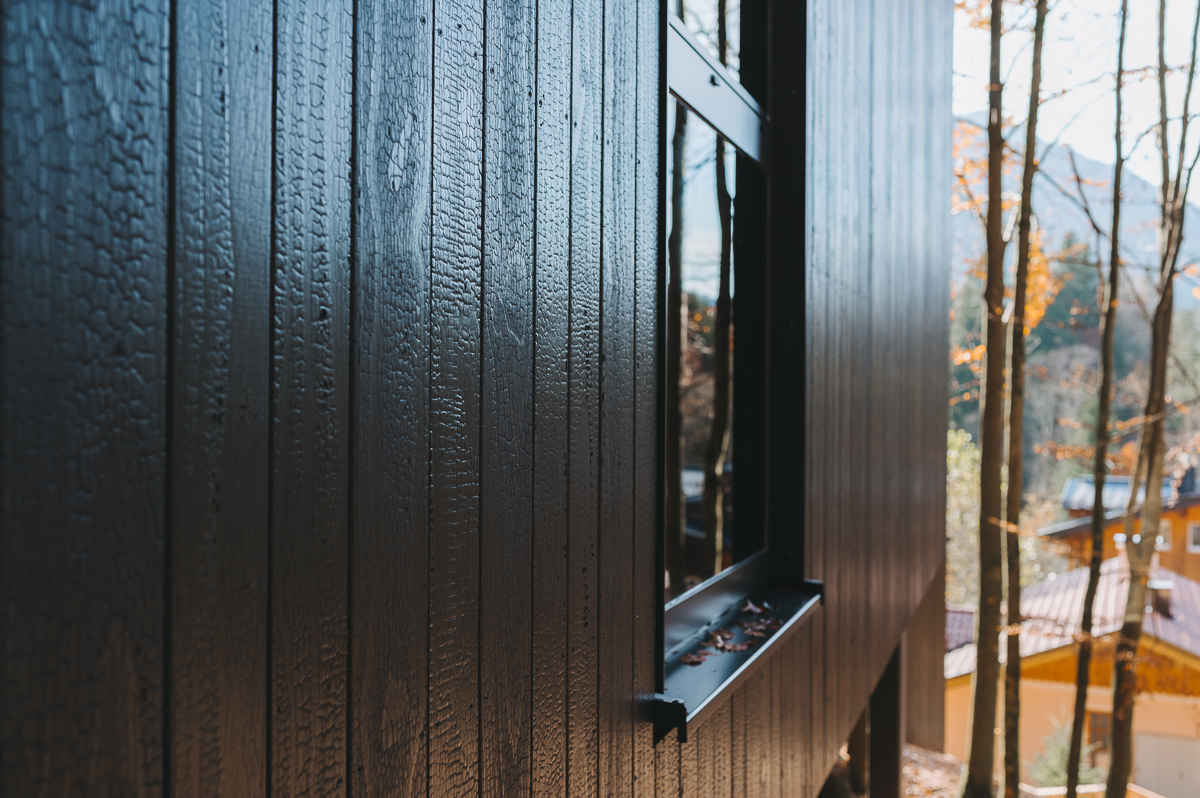 Detailaufnahme einer dunklen, verkohlten Kebony-Holzfassade am Chalet im Naturhotel Fischerwirt Salzburg. Im Hintergrund herbstlicher Wald und Berglandschaft.
