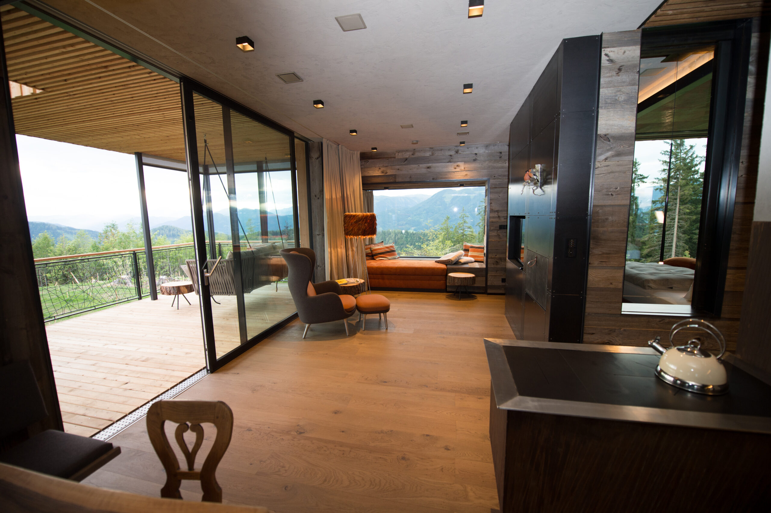 Gemütlicher Wohnraum mit Panoramafenstern, Holzverkleidung aus Mareiner Piz Bernina, großem Loungesessel und Fensterbank-Sofa mit Blick auf die Berge, angrenzend an eine überdachte Holzterrasse.