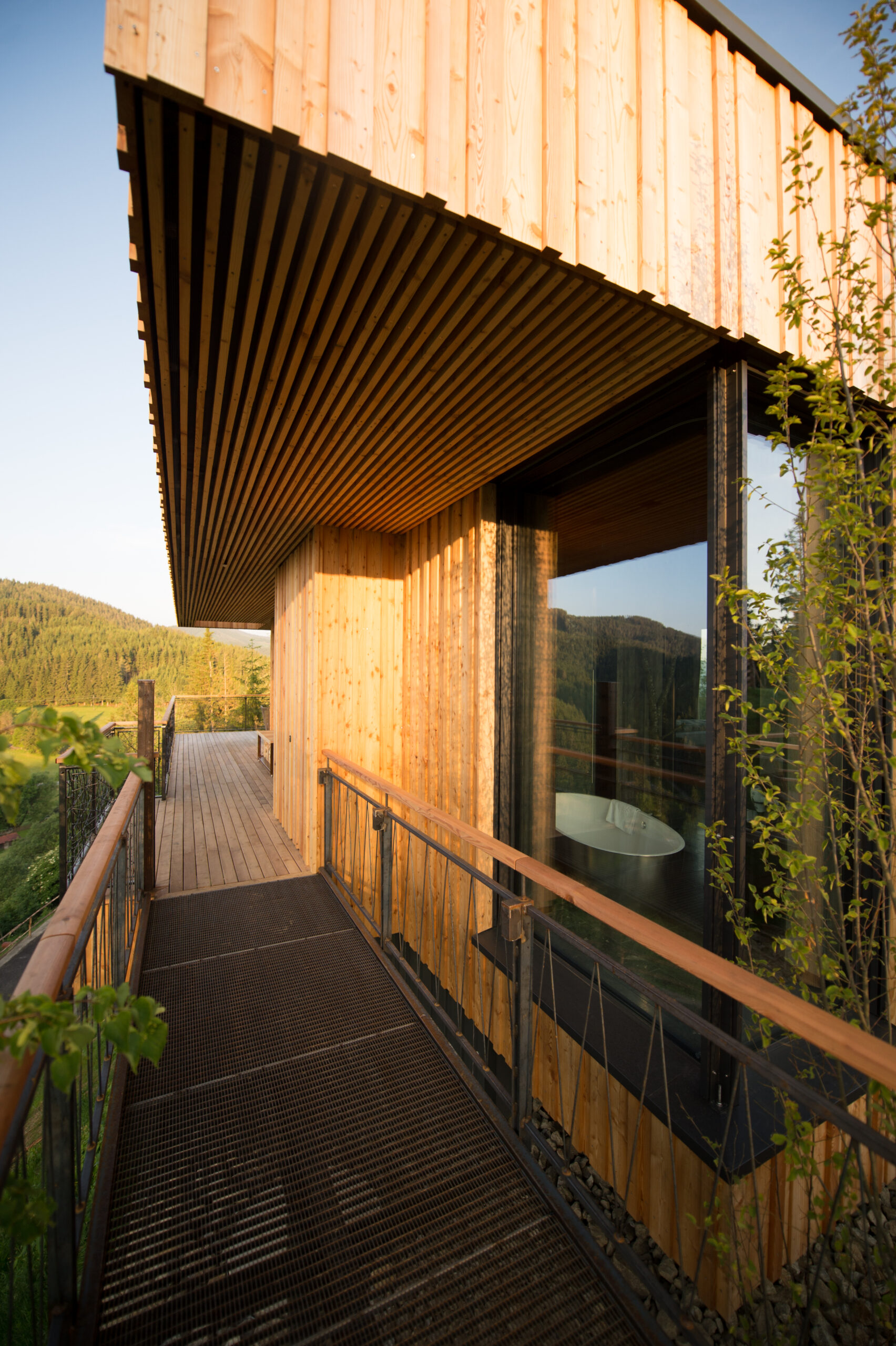 Holzsteg führt entlang einer modernen Chalet-Fassade aus vertikalem Mareiner Holz Piz Bernina, flankiert von Glasfront und Blick auf bewaldete Hügel im Abendlicht.