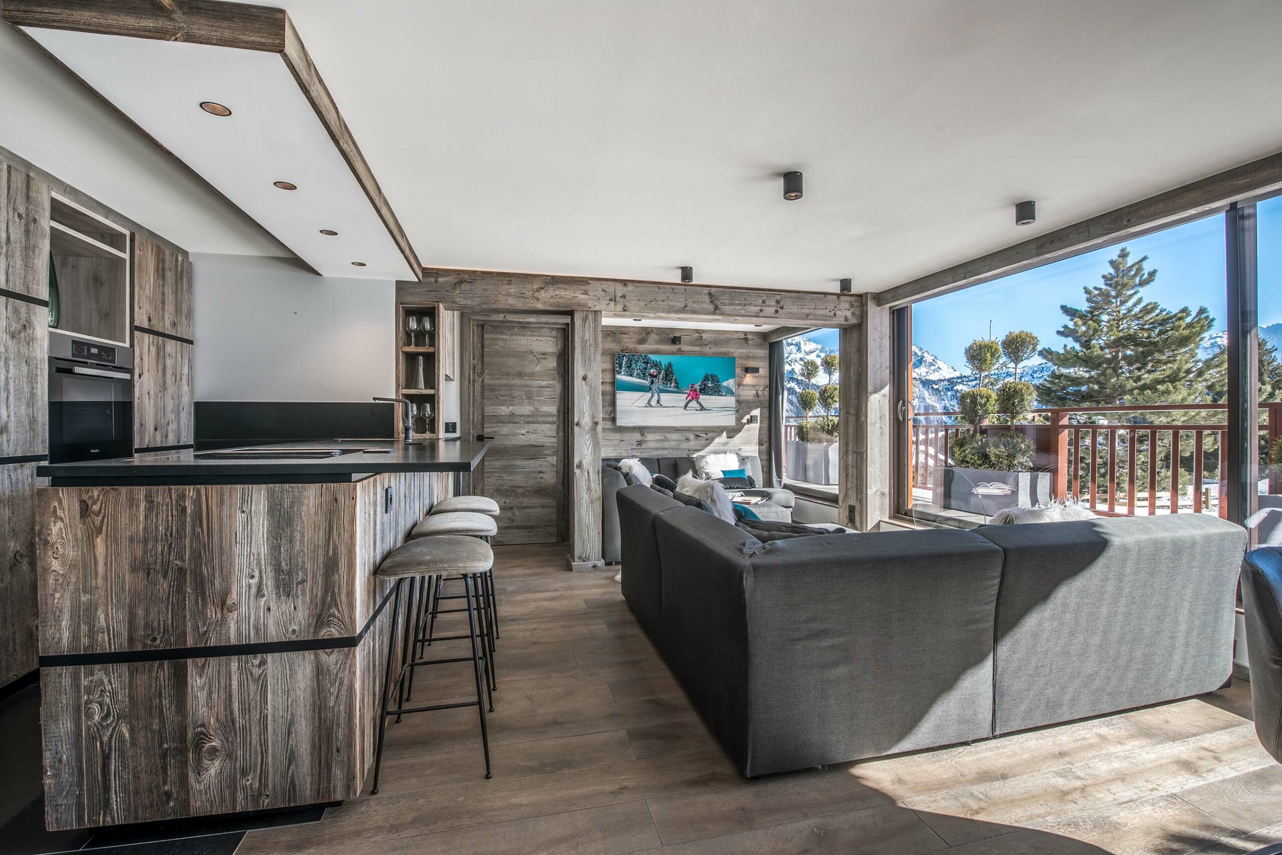 Großzügige Wohnküche mit Küchenzeile und Barbereich aus Mareiner Altholz Zermatt Gris, kombiniert mit moderner Einrichtung und Panoramablick auf die verschneiten Alpen im Hotel Paradis du Pralong.