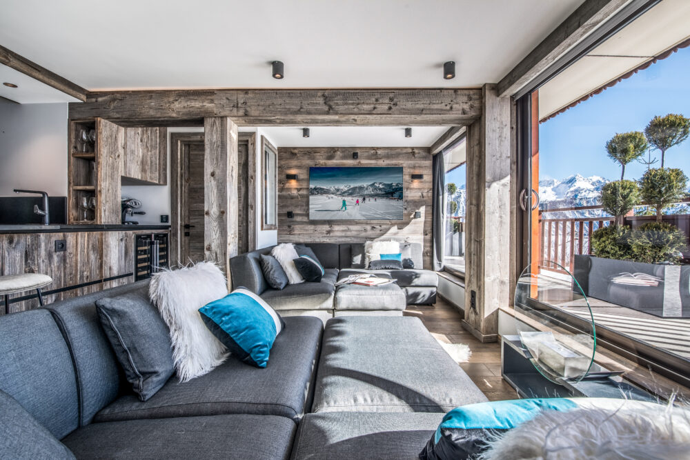 Modernes Wohnzimmer mit grauem Sofa und Altholz-Wänden aus Mareiner Zermatt Gris. Große Fenster bieten freien Blick auf verschneite Berge. Stilvolle Kombination aus rustikalem Holz und elegantem Design.